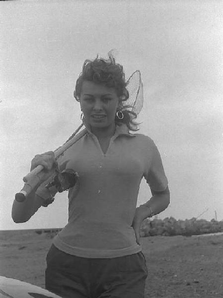 Località non identificata. Sofia Loren sulla spiaggia. L'attrice tiene in mano una canna e un retino da pesca
