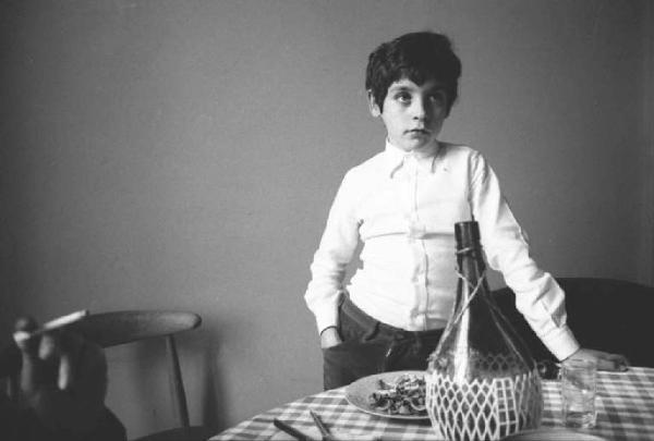 Ritratto maschile - bambino in piedi dietro un tavolo sul quale è appoggiato un fiasco di vino
