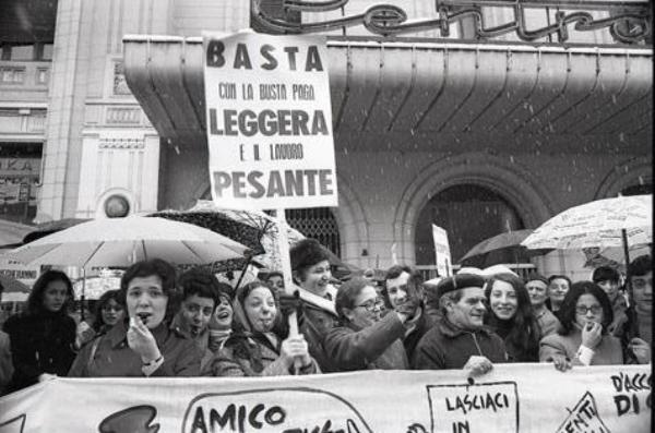 Sciopero dei Tessili di Monza: primo piano di alcuni lavoratori. Uno dei cartelli mostra la scritta "Basta con la busta paga leggera e il lavoro pesante"
