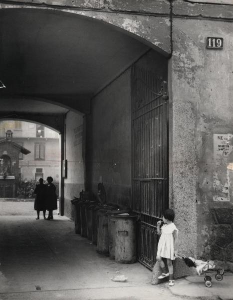 Milano - Quartiere Lampugnano - Ingresso di case di ringhiera - Due donne vicino ai bidoni dei rifiuti - Bambina con gioco