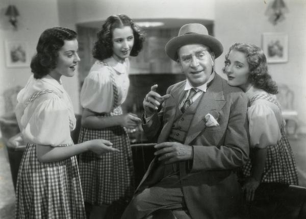 Scena del film "Una famiglia impossibile" - Regia Bragaglia, Carlo Ludovico, 1940 - Armando Falconi, con la mano sinistra in aria, guarda verso destra circondato da tre ragazze del Trio Primavera.