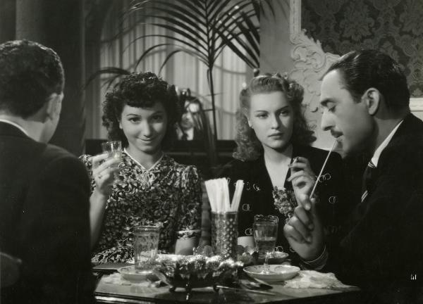 Scena del film "Fari nella nebbia" - Regia Franciolini, Gianni, 1942 - Mariella Lotti, con una sigaretta in mano, osserva Carlo Lombardi che si porta uno stecco alla bocca. A sinistra, due attori non identificati. Lei guarda verso l'obbiettivo.