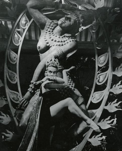 Scena del film "La figlia di Mata Hari" - Merusi, Renzo, 1954 - Un'attrice non identificata, in abiti indiani, è in posa di danza con la gamba destra sollevata e le braccia incrociate all'altezza del viso mentre guarda in alto a sinistra.
