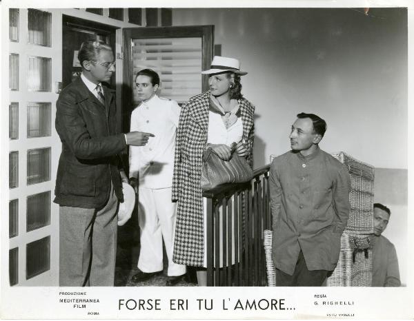Scena del film "Forse eri tu l'amore" - Righelli, Gennaro, 1939 - Loretta Vinci, appoggiando la sua borsa alla ringhiera, si rivolge con lo sguardo a Romolo Costa che la indica. Dietro, tre attori non identificati. Uno di loro è in divisa.
