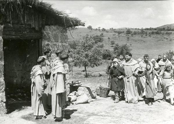 Scena del film "Francesco, giullare di Dio" - Rossellini, Roberto, 1950 - A destra, un gruppo di frati e pastorelli, mano nella mano, viene accolto da quattro attori non identificati in veste da frate, a sinistra. Dietro di loro, un casale.
