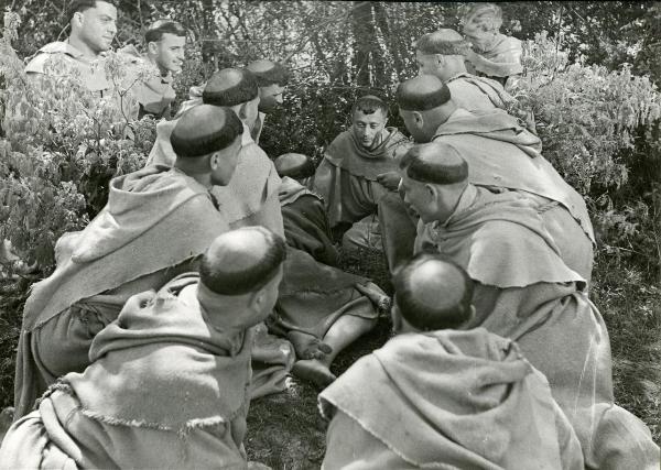 Scena del film "Francesco, giullare di Dio" - Rossellini, Roberto, 1950 - In mezzo alle sterpaglie, al centro, Nazario Gerardi è attorniato da un gruppo di frati di spalle che lo ascoltano.