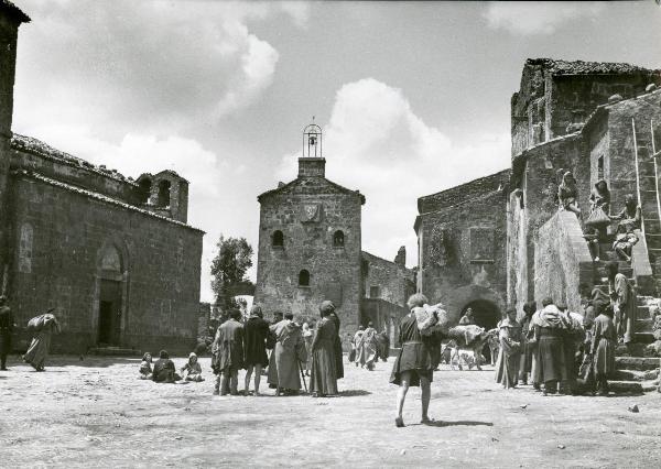 Scena del film "Francesco, giullare di Dio" - Rossellini, Roberto, 1950 - Al centro di una piazza, molti attori non identificati sono ritratti durante la loro vita quotidiana.
