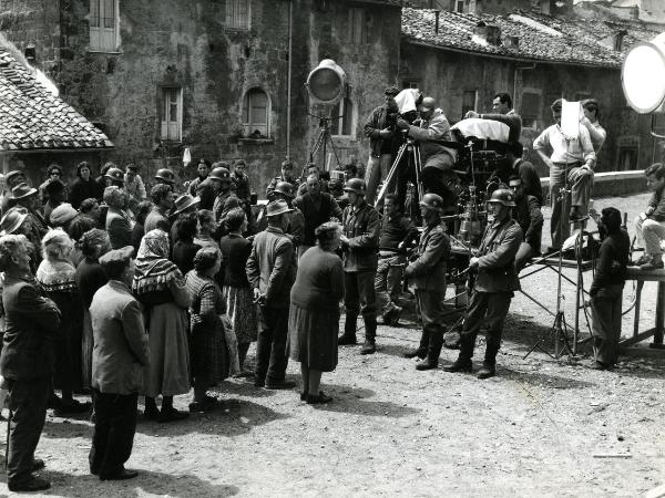 Fotografia sul set di "Un giorno da leoni" - Loy, Nanni, 1961 - Su un impalcatura, alcuni operatori con Nanni Loy, a sinistra, e Marcello Gatti, al centro, riprendono la scena: alcuni soldati tengono in riga una folla di attori non identificati.