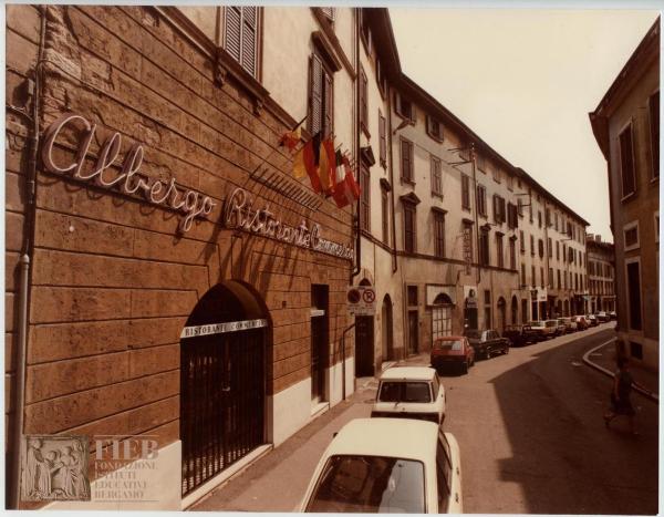 Albergo Commercio - Bergamo - Complesso di Santo Spirito - Esterno: via Torquato Tasso - Automobili parcheggiate - Fiat 850 - Renault r5 - Una donna attraversa la strada - Bandiere - Negozi