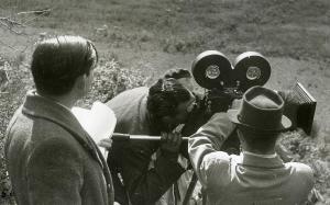 Fotografia sul set di "Un garibaldino al convento" - De Sica, Vittorio, 1942 - Al centro, il regista Vittorio De Sica guarda all'interno della cinepresa. Ai lati, due operatori non identificati di spalle.