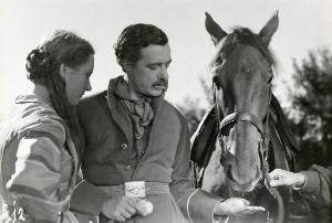 Fotografia sul set di "Un garibaldino al convento" - De Sica, Vittorio, 1942 - Al centro, il regista Vittorio De Sica dà da mangiare con la mano sinistra a un cavallo posizionato a destra. A sinistra, Carla Del Poggio di profilo, osserva la scena.