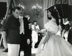 Fotografia sul set di "Il gattopardo" - Visconti, Luchino, 1963 - A destra, Claudia Cardinale in abito elegante guarda Burt Lancaster a sinistra. Davanti a Lancaster, il regista Luchino Visconti guarda verso il basso.