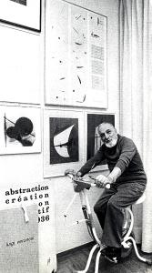 Fotografia sul set di "Un giorno alla Olivetti" - Veronesi, Luigi, 1947 - In uno studio con appesi dei quadri, Luigi Veronesi sulla ciclette guarda verso la cinepresa.