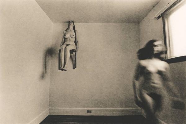 Coppia di donne nude in una stanza vuota; una è seduta e appesa al soffitto