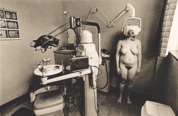 Donna nuda in piedi in uno studio odontoiatrico; le copre il viso uno strumento medico