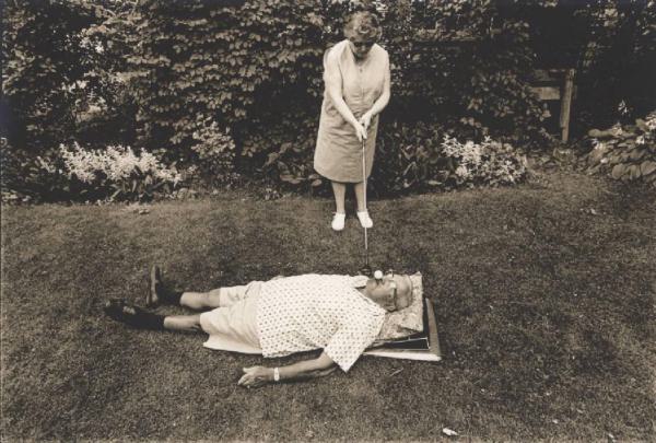 Donna anziana colpisce pallina da golf appoggiata sul volto di uomo anziano sdraiato in giardino