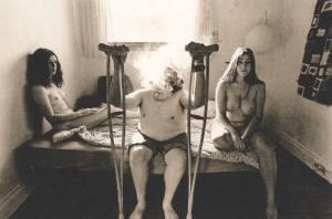Uomo con stampelle siede sul letto tra due ragazze nude