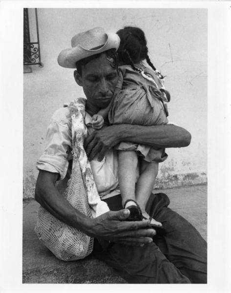 El Salvador. Chalatenango. Ritratto. Un uomo seduto tiene in braccio una bambina. Cappello