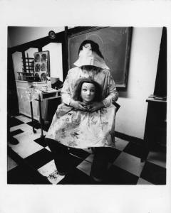 Donna seduta con il viso coperto e una testa con parrucca in grembo - Capelli a terra - Sulla parte una lavagna - Sullo sfondo un poster con bigodini