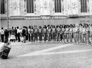 Piazza Duomo: La foto. Milano - Piazza del Duomo - Gruppo di atlete posa davanti ad un uomo accovacciato con macchina fotografica. Tuta da ginnastica. In secondo piano sono visibili numerose persone. Sullo sfondo parte del Duomo