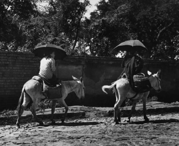 Coppia di donne contadine sopra due asini nei pressi di un muro di cinta