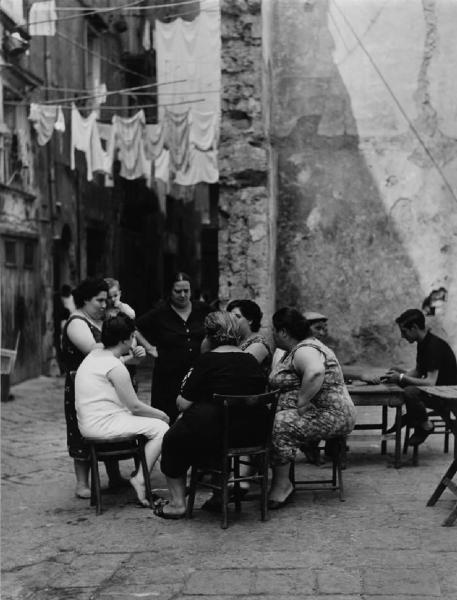 Napoli: Conversazioni. Napoli - Vicoli - Donne sedute in cerchio su sedie e altre in piedi - Sullo sfondo panni stesi e due ragazzi seduti a un tavolo