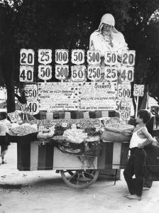 Napoli: Si vende si compra/ Commercio. Napoli - Carretto venditore ambulante di frutta secca, arachidi: bambino commerciante - Monumento, statua sullo sfondo