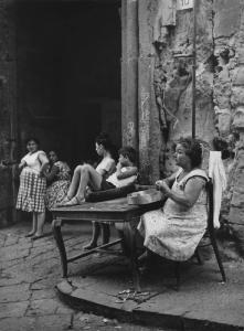 Napoli: Nei Bassi. Napoli - Vicoli - Donna pela patate a un tavolo e bambino seduto sul tavolo dentro una ruota