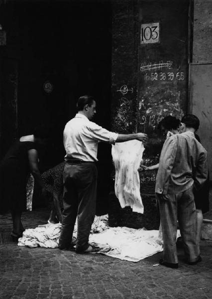 Napoli: Commercio. Napoli - Strada - Venditore ambulante di indumenti per donna: uomo - Uomini e donne - Scritte sul muro