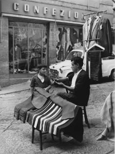 Napoli: Commercio. Napoli - Vicoli - Banco di abiti da uomo - Venditore ambulante seduto su una sedia, bambino