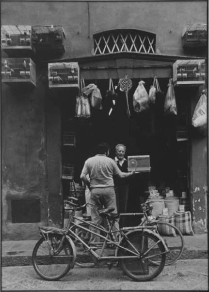 Firenze - Negozio visto dalla strada: gabbie con uccellini appese, sacchi di legumi, battipanni, sacchi - Due uomini sull'ingresso: Arnoldo M., civaiolo e cliente