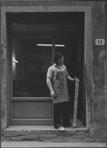 Firenze - Bottega vista dalla strada: vetrina - Ritratto maschile a figura intera: Andrea O., intagliatore con pezzo di legno intagliato