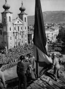 Gorizia. Militari innalzano bandiera italiana sul tetto di un edificio. Folla radunata sulla piazza di una chiesa