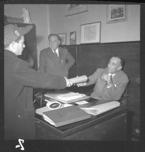 Mantova (?) - Ufficio Turismo (?) - Un uomo con cappello nell'atto di consegnare un carteggio ad un altro seduto alla scrivania - Un uomo in piedi