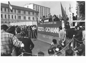 Celebrazione del 30° anniversario della Liberazione - Comizio in piazza - Palco - Giovanni Pesce al microfono - Operai con tuta da lavoro - Bandiere - Striscione - Gonfalone