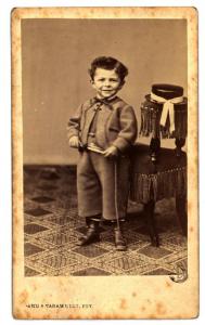 Ritratto infantile - Bambino in piedi accanto a una poltroncina con frange