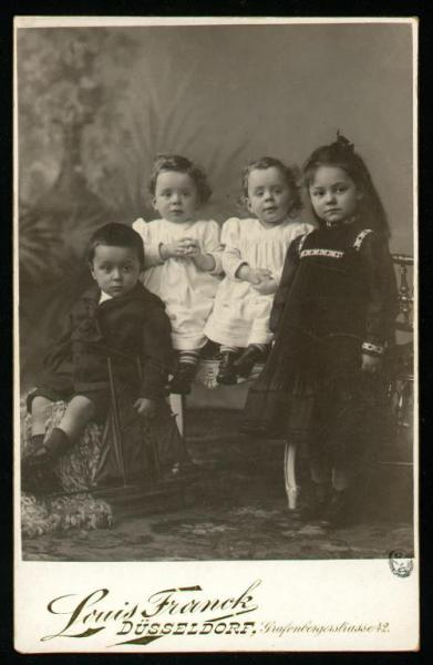 Ritratto di famiglia - Quattro bambini, una bambina in piedi, un bambino seduto e due gemelle in abito chiaro sedute su divanetto