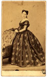 Ritratto femminile - Donna in abito decorato con motivi geometrici, in piedi con le mani appoggiate allo schienale di una poltrona