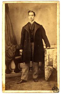 Ritratto maschile - Uomo con pantaloni a quadri e cappello a tuba, in piedi appoggiato a un tavol