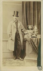 Ritratto maschile - Uomo in piedi con cappello a tuba accanto a un tavolo con coppe