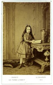 Ritratto infantile - Bambina in piedi, vicino a un tavolo, davanti ad un libro aperto