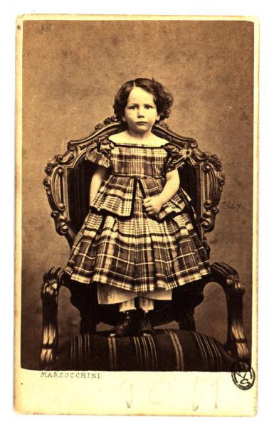 Ritratto infantile - Bambina in abito a quadri in piedi su una poltrona
