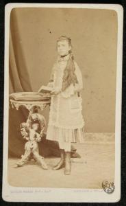 Ritratto femminile - Giovane dai lunghi capelli e con stivaletti in piedi accanto a un tavolino