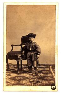 Ritratto infantile - Bambino con cappello, in piedi accanto a una poltroncina