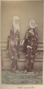 Scena di genere - Due donne giapponesi, in piedi, indossano, pur essendo in un interno, il copricapo per il freddo e si tengono mano nella mano - Per terra un tappeto