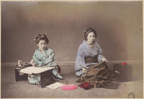 Ritratto di gruppo - Due giovani donne giapponesi, in kimono, sedute sui talloni - Una intenta a scrivere su un tavolino - L'altra a cuicire - Al centro un ventaglio posato per terra