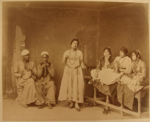Ritratto di gruppo - Una donna danza accompagnata da due suonatori uomini e da una suonatrice di tamburello - Una donna seduta la guarda a gambe incrociate e un'altra fuma guardando verso l'obiettivo