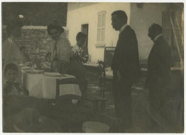 Esterno, cortile di una casa - Gruppo di uomini, donne e bambini attorno a una tavola imbandita - Pasto