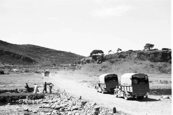 Viaggio in Africa. Paesaggio africano: due autocarri militari italiani percorrono una strada sterrata che si snoda tra colline rocciose punteggiate di alberi isolati