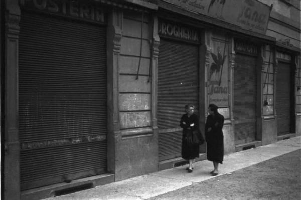 Donne passeggiano per una via cittadina semideserta - serrande dei negozi abbassate